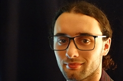 [ai] A young man with glasses and long hair tied back, smiling at the camera. He is standing in front of a dark background, wearing a checkered shirt.